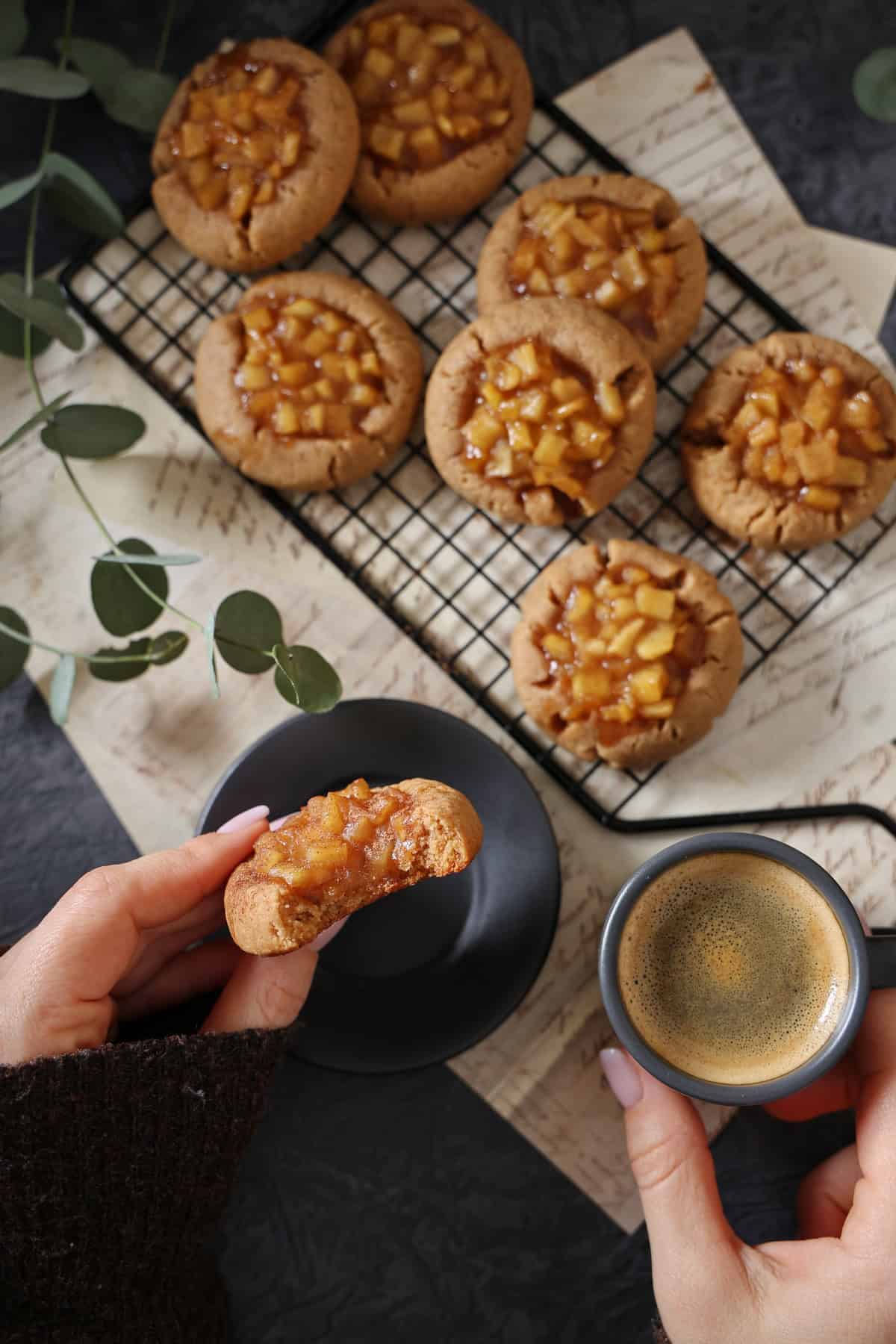 Two hands holding an apple cinnamon cookie and a small cup of coffee. Above it, a black baking sheet with apple cinnamon cookies.