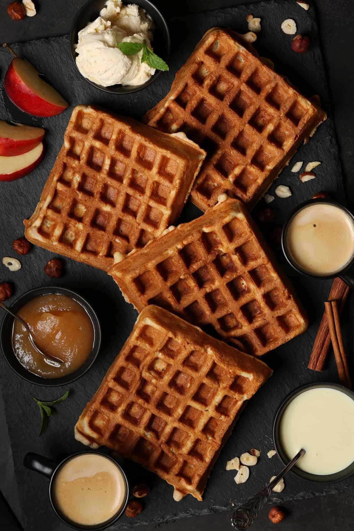 Several apple nut waffles on a dark background. Next to them are small bowls of applesauce and vanilla sauce.