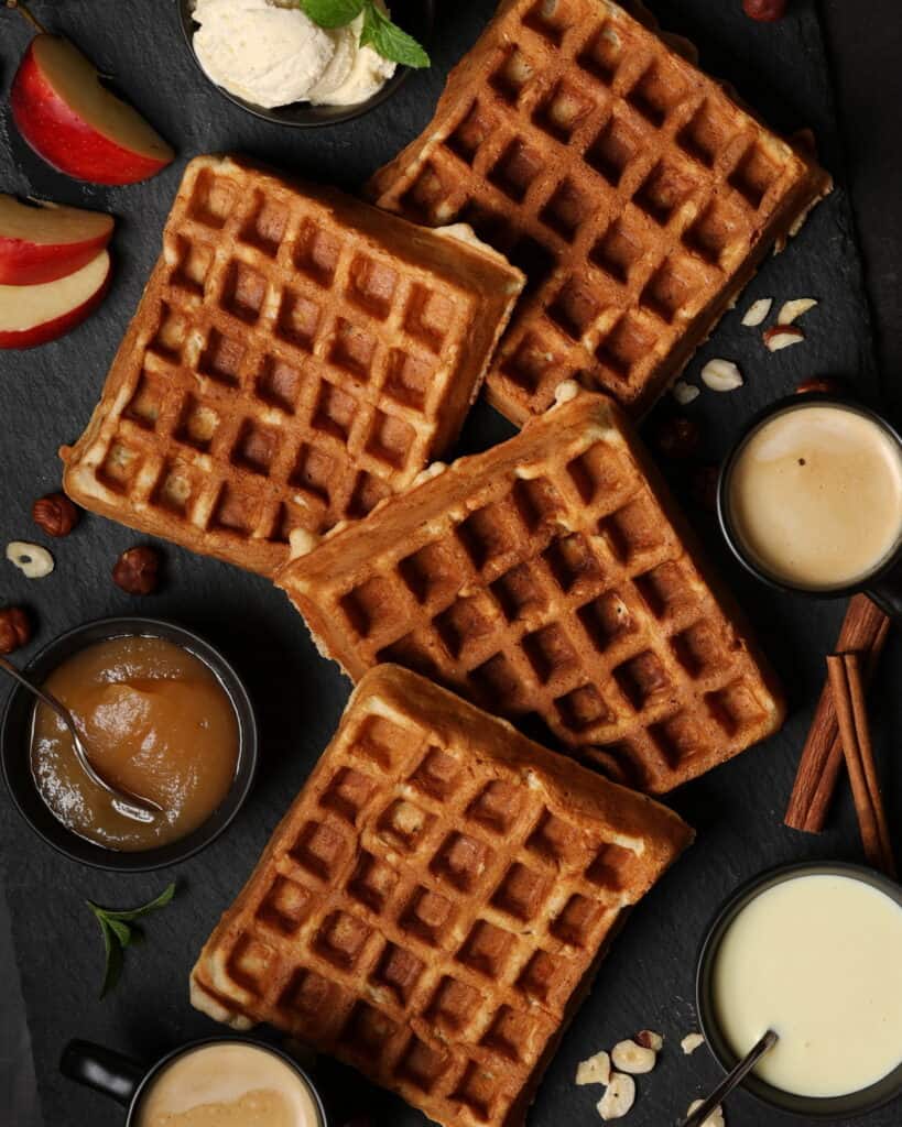Several apple nut waffles on a dark background. Next to them are small bowls with applesauce and vanilla sauce.