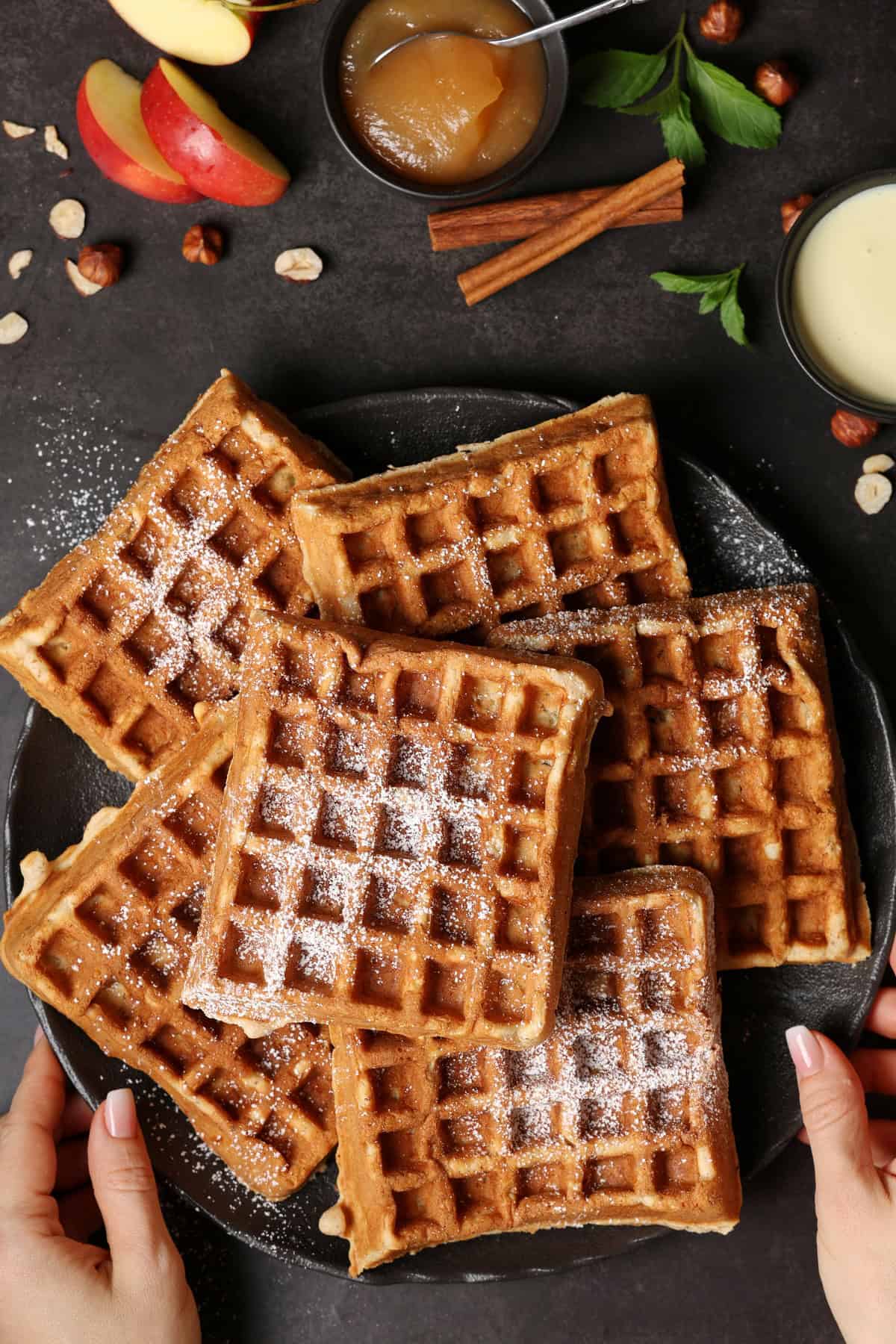 Two hands holding a dark plate with apple nut waffles.