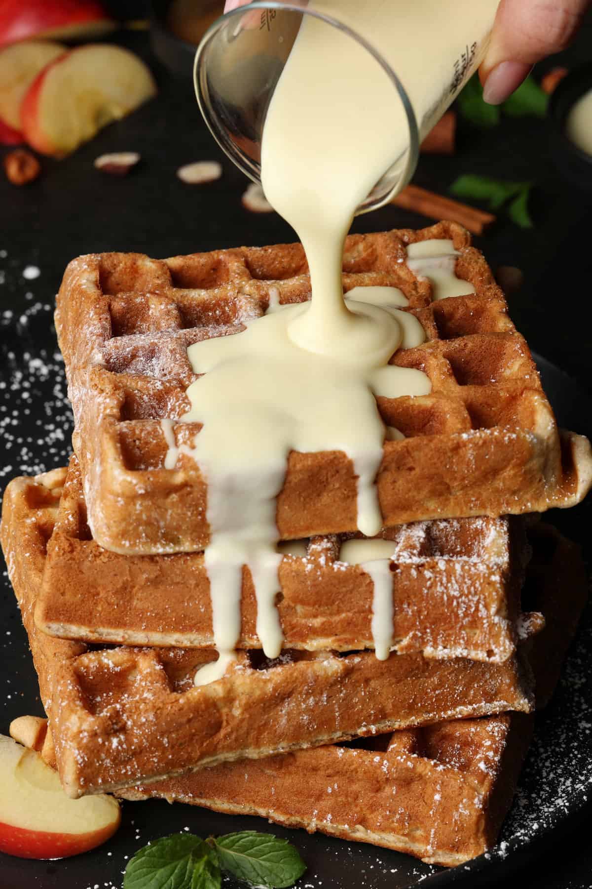 A hand pouring vanilla sauce over a stack of apple nut waffles.