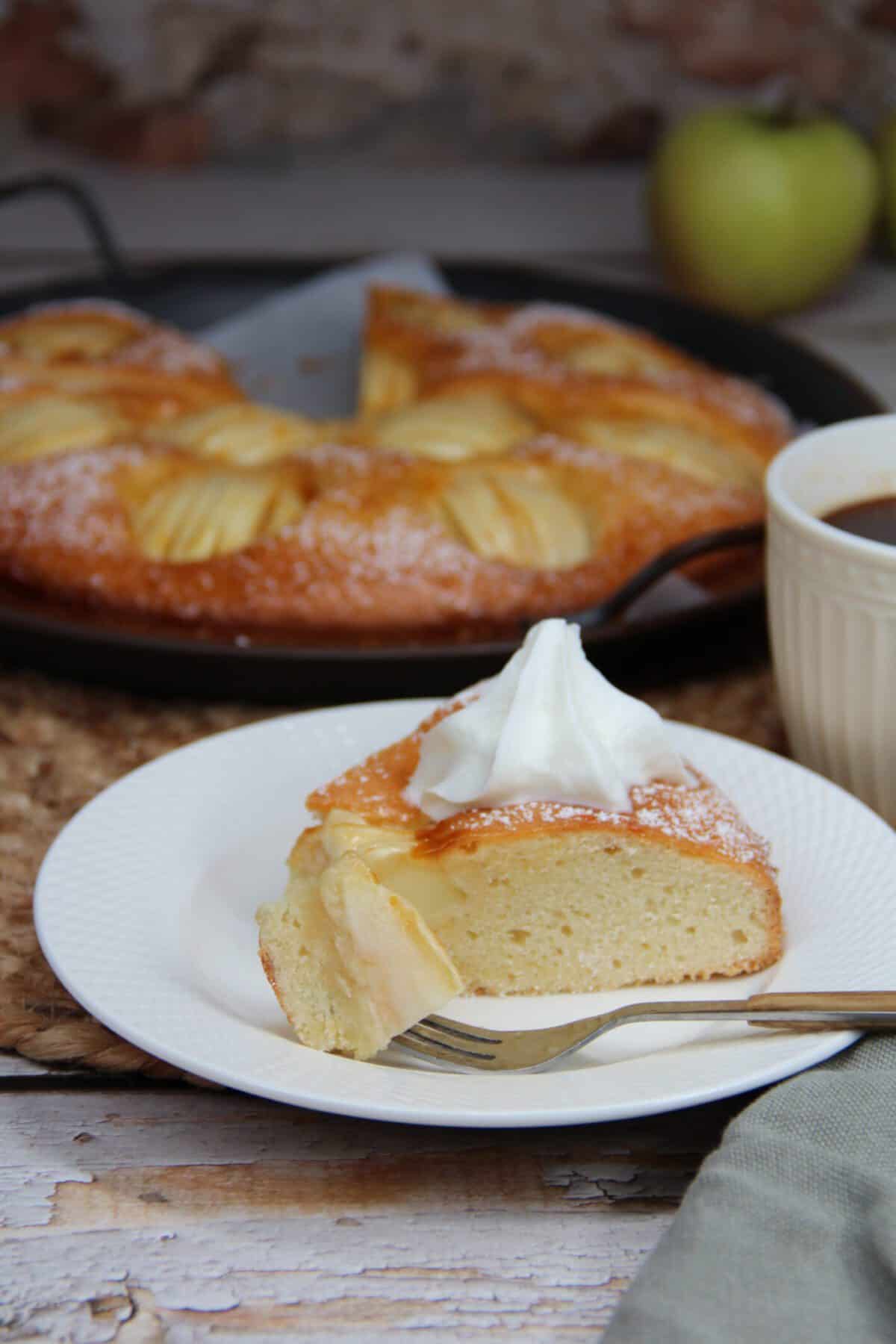 A white plate with a slice of apple cake served with whipped cream.