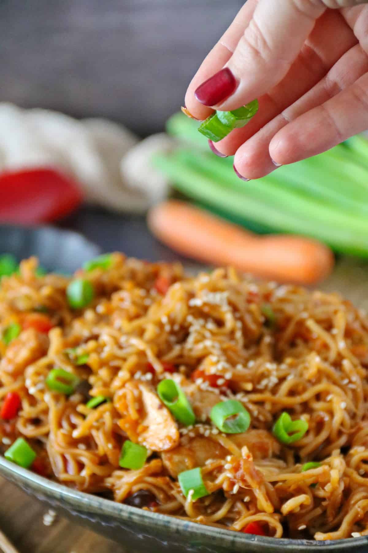 A hand sprinkling sliced green onions over a bowl of Asian noodles with chicken.
