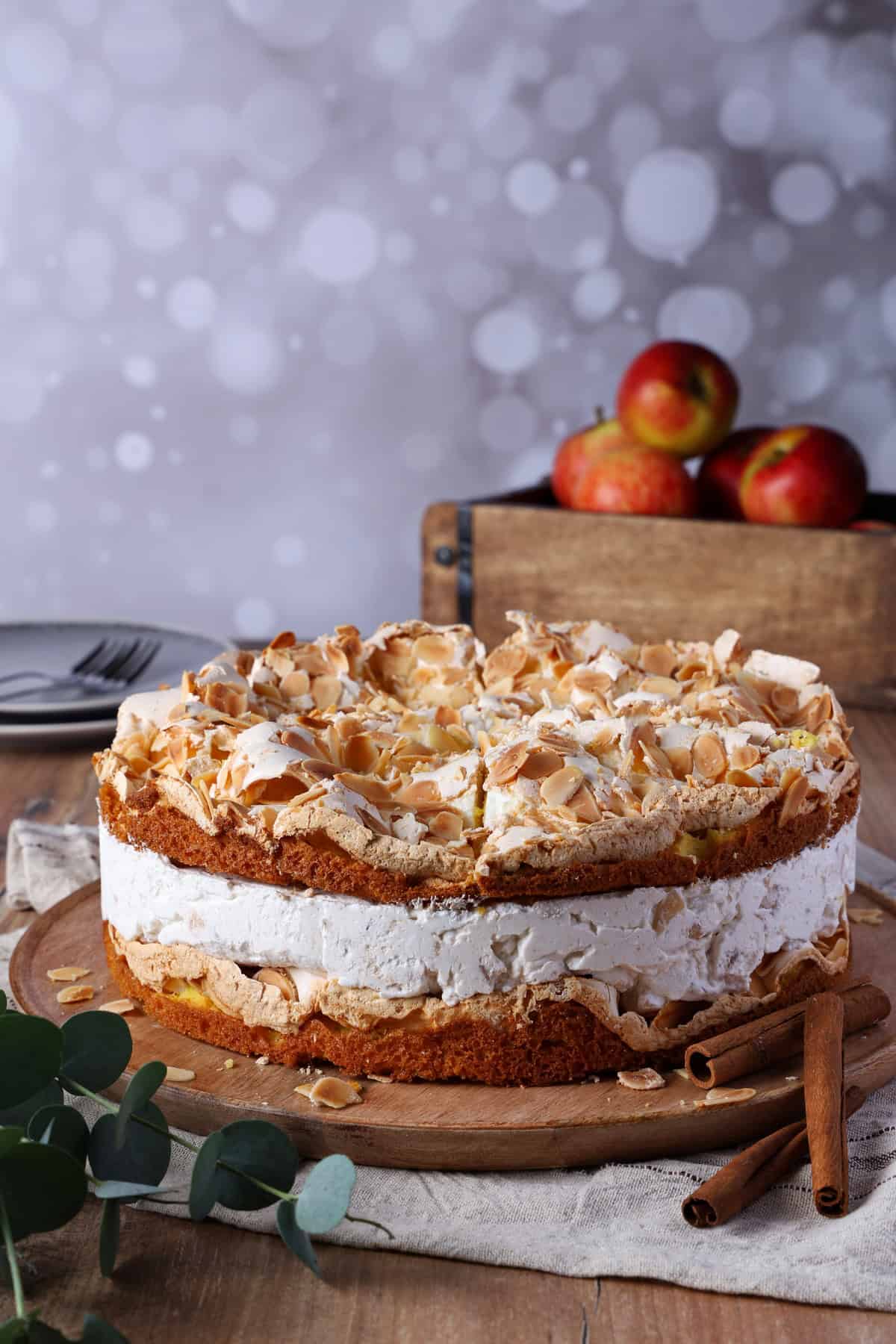 A baked apple meringue cake on a round wooden serving tray. Behind it is a wooden crate filled with apples.