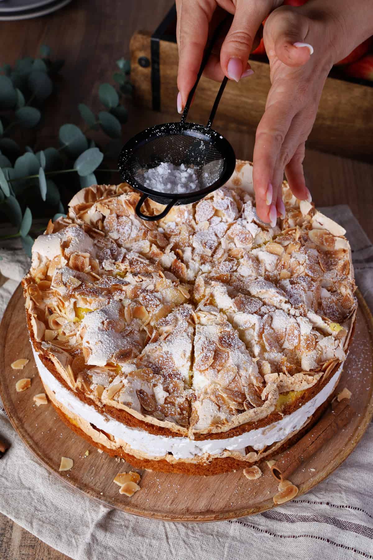 Two hands dusting powdered sugar through a small fine-mesh strainer over a baked apple meringue cake.