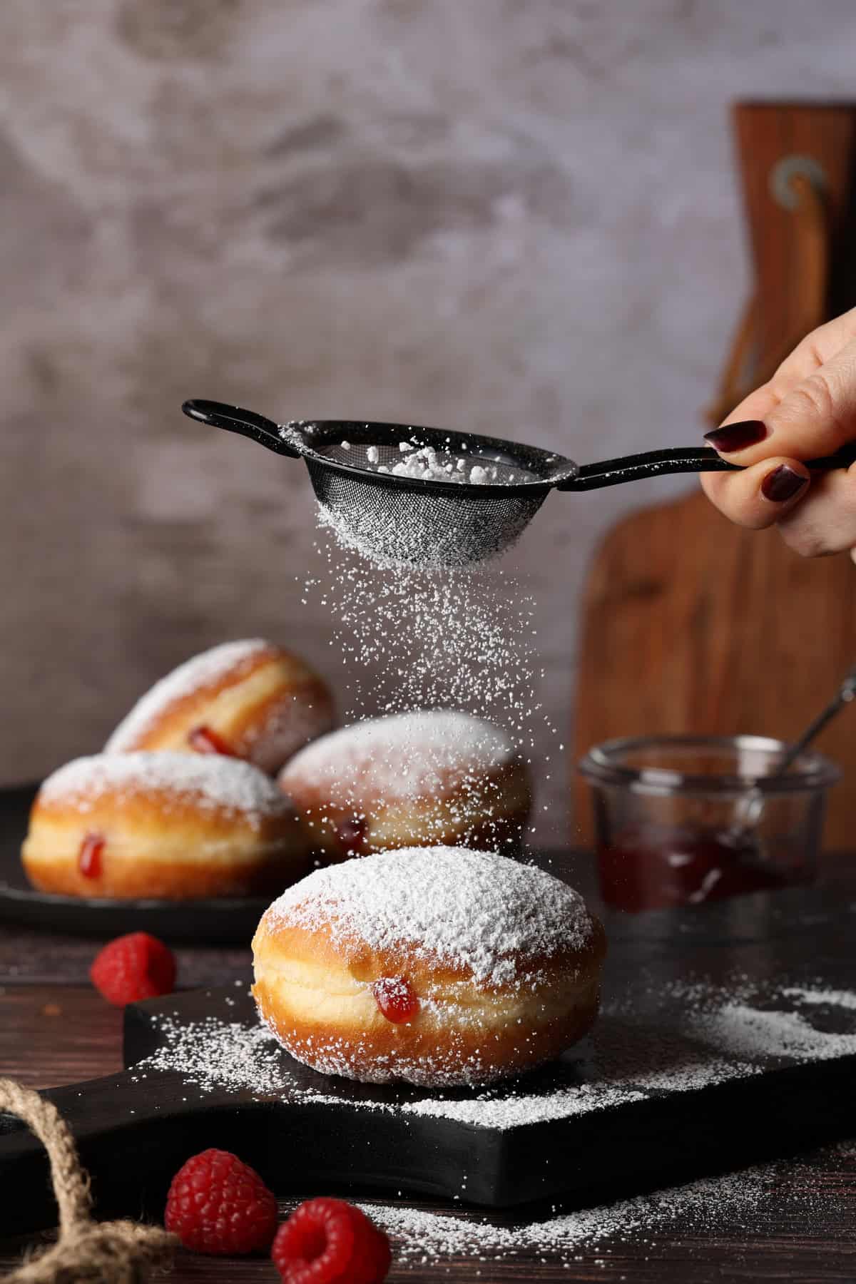 A hand dusting powdered sugar from a small fine mesh strainer over a jelly donut. More donuts in the background.