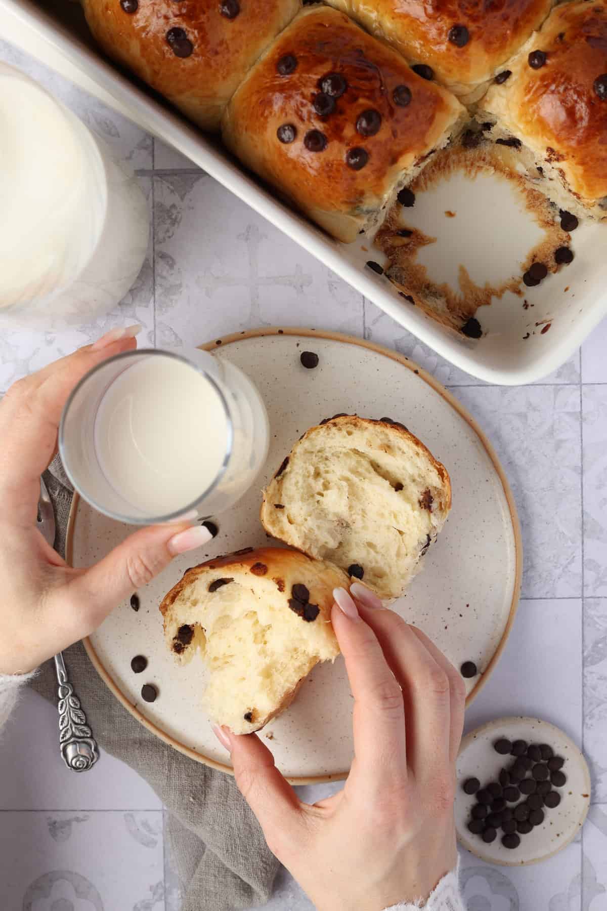 Two hands holding a glass of milk and a chocolate roll on a plate. Above it, part of a baking dish with more chocolate rolls.