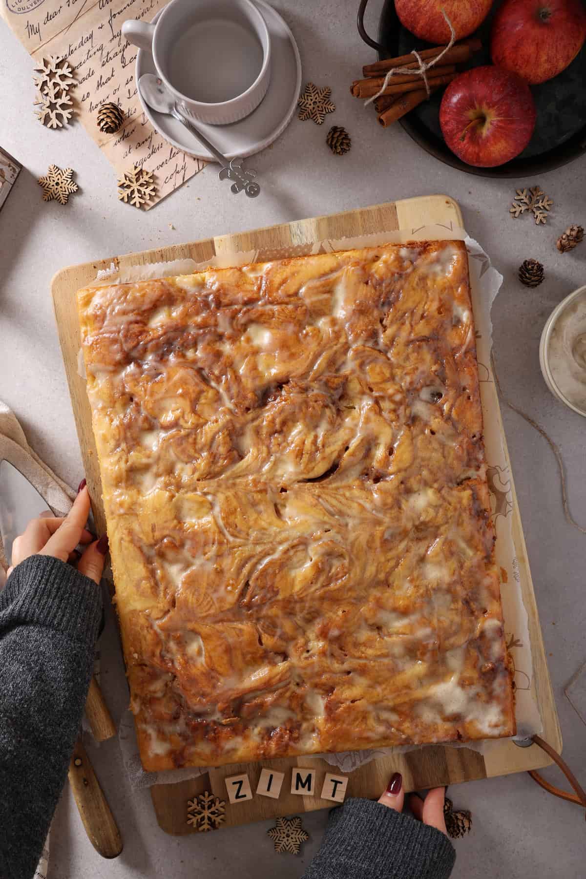 Two hands holding a wooden cutting board with a whole apple cinnamon roll cake.