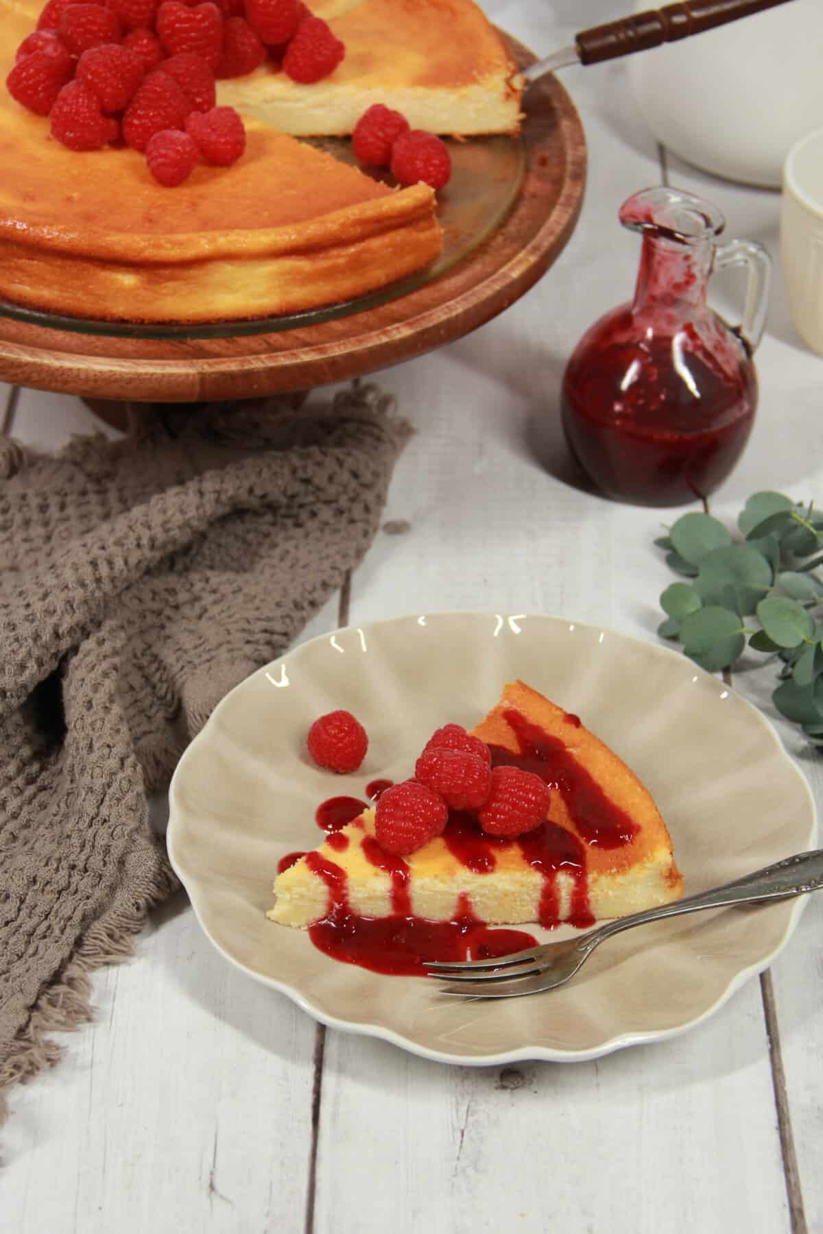 A slice of crustless cheesecake with raspberries on a plate, with the rest of the cheesecake on a serving platter in the background.
