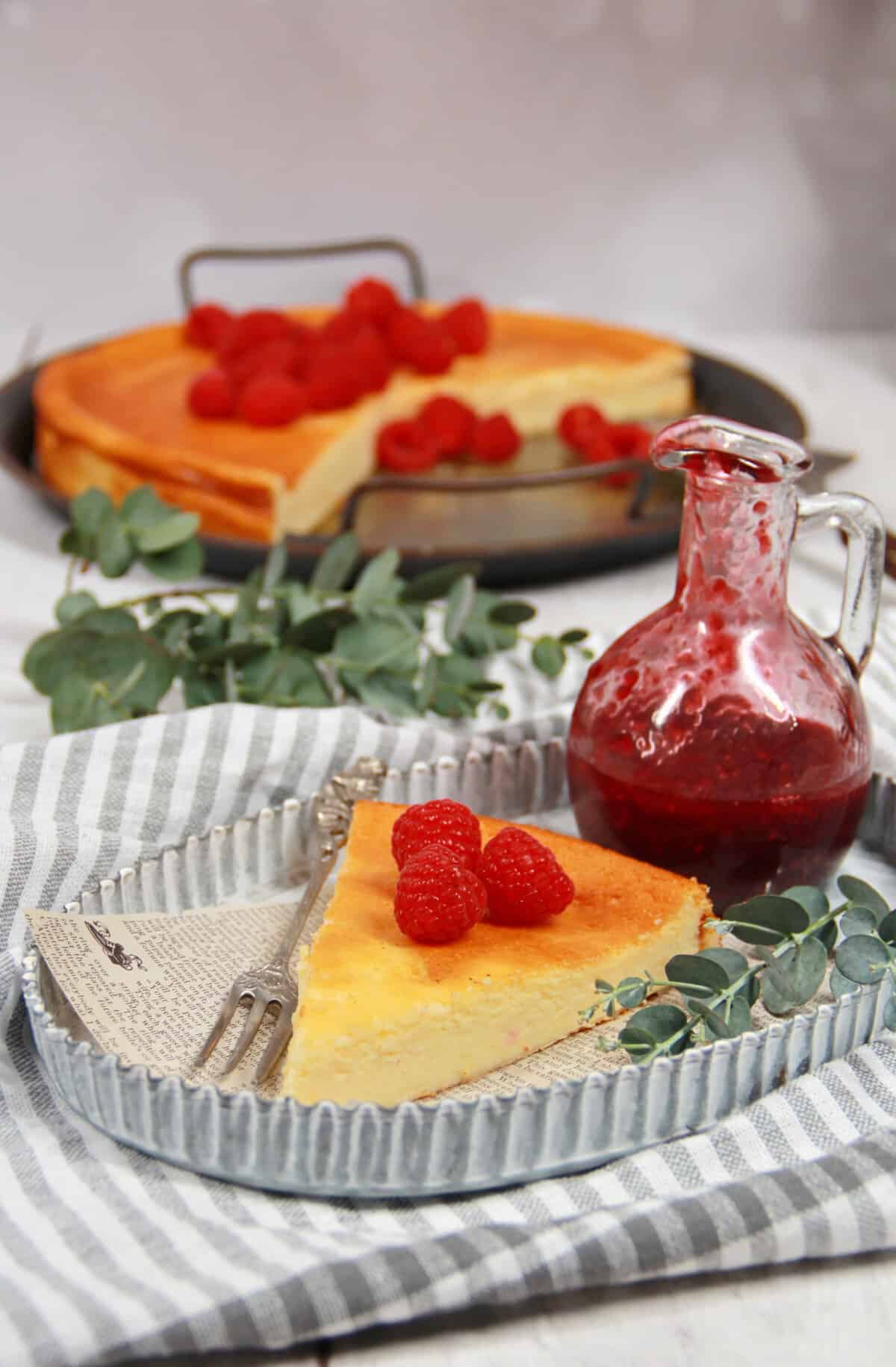A slice of crustless cheesecake with raspberries on a plate, with the whole cheesecake on a serving platter in the background.