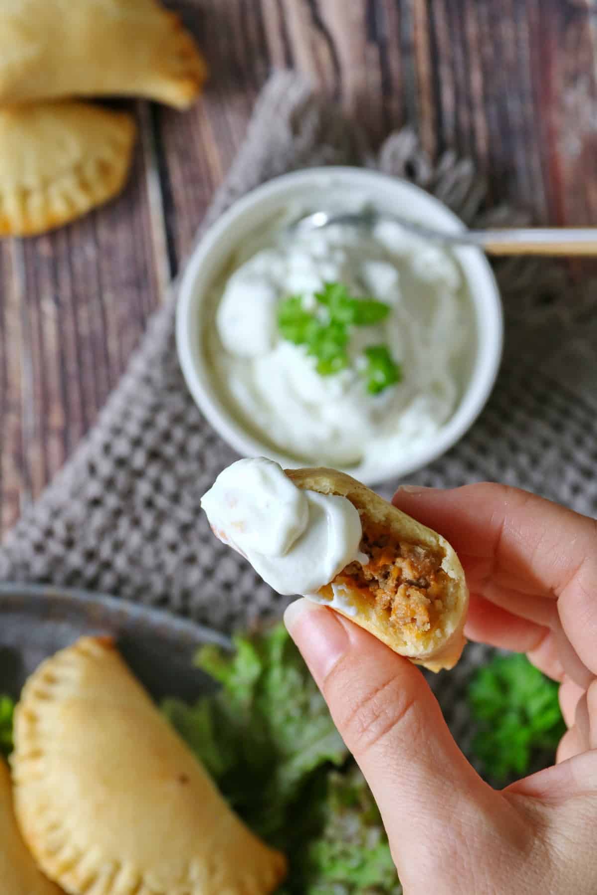 A hand holding a piece of empanada dipped in sauce, with more empanadas and a small bowl of dip in the background.