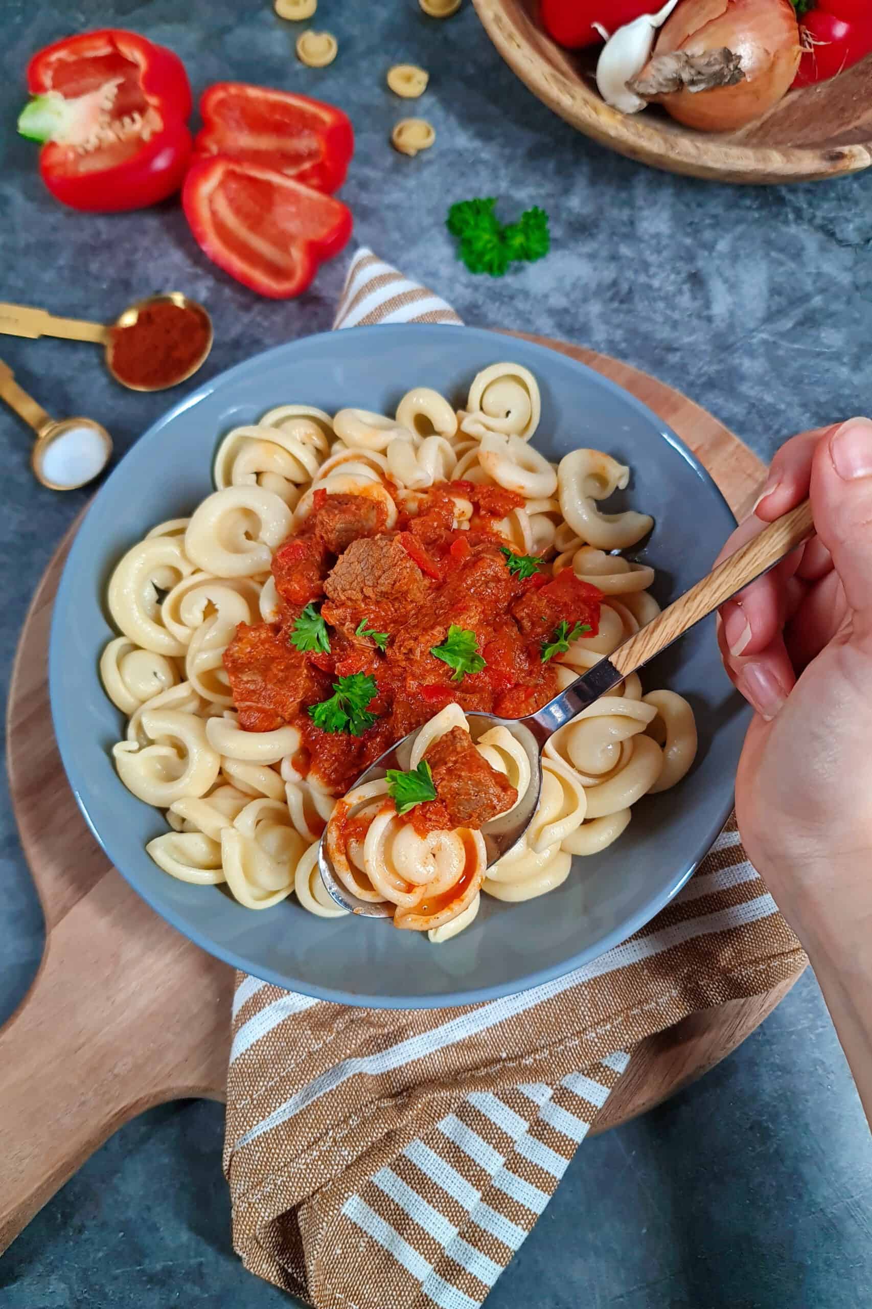 A hand holding a spoon scoops beef goulash and noodles from a plate.