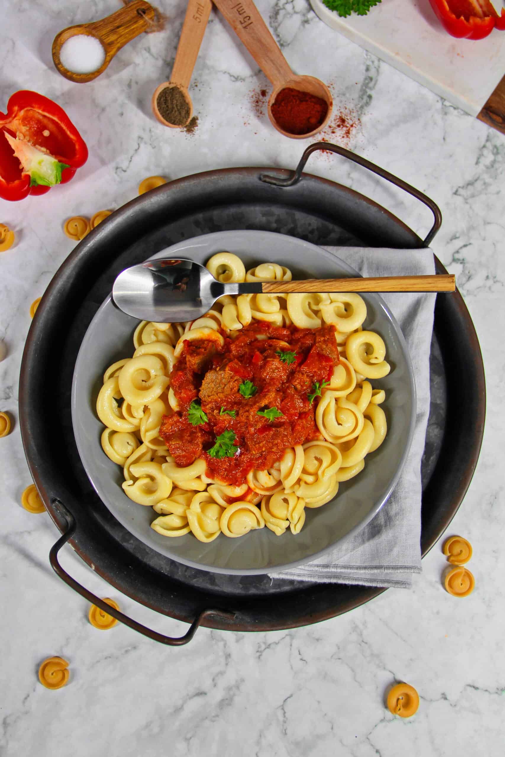 A deep gray bowl filled with beef goulash, bell peppers, and noodles on a round serving tray.