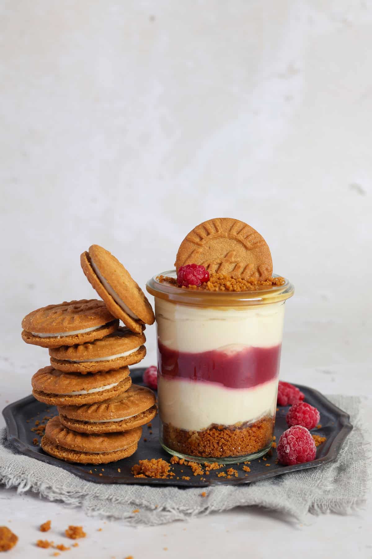 A glass of Lotus Biscoff dessert with raspberries on a small plate. Next to it, a stack of Lotus Biscoff cookies and fresh raspberries.