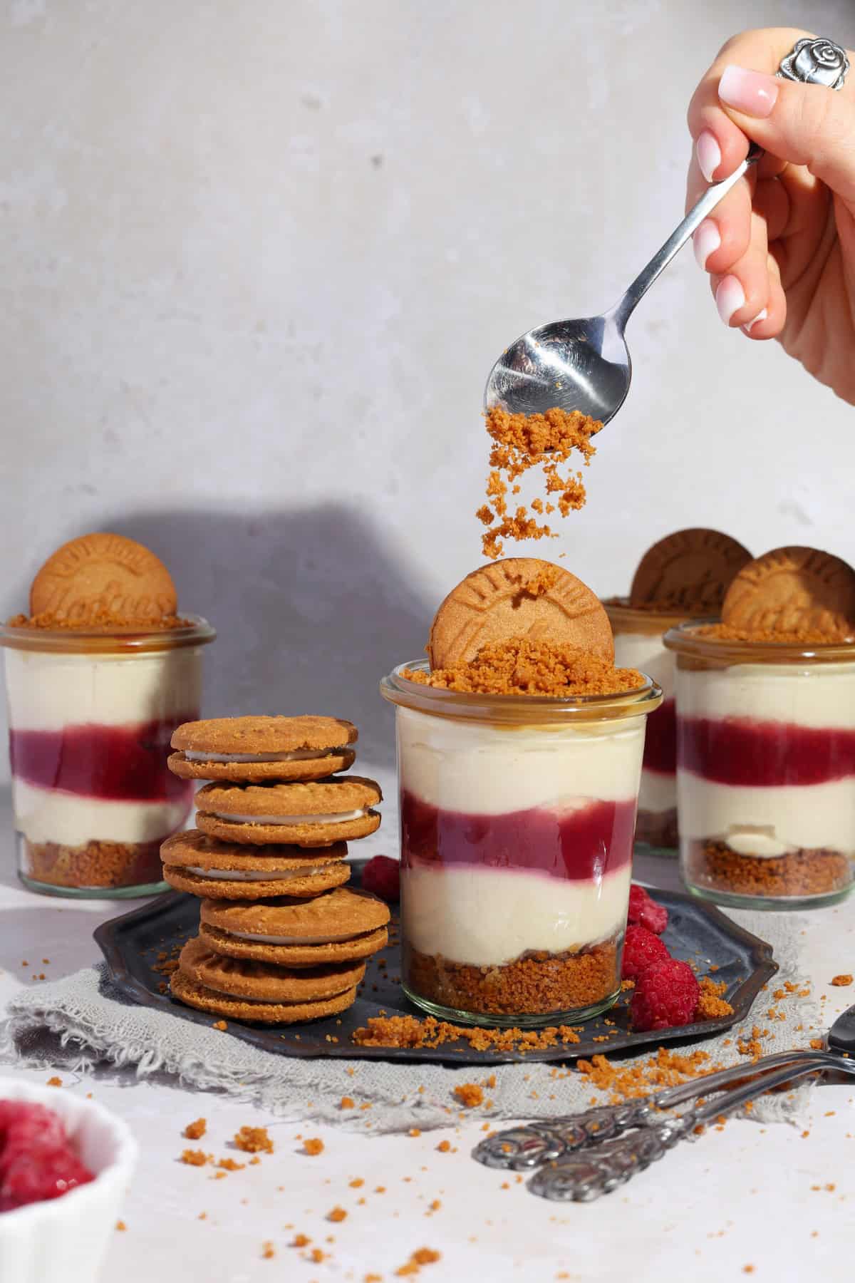 Several glasses of Raspberry Lotus Biscoff dessert cups. A hand is sprinkling cookie crumbs over one of the glasses with a spoon.