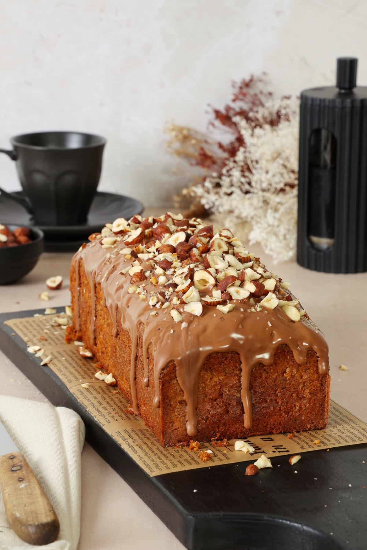 A moist nut cake with chocolate glaze and chopped hazelnuts on a dark serving board.