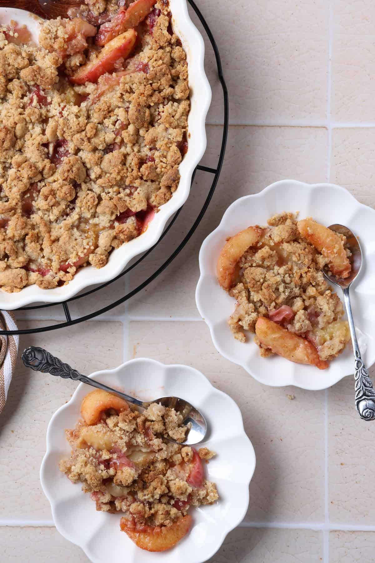 Two small plates of peach crumble with hazelnut streusel topping, with a baking dish of the remaining crumble in the background.