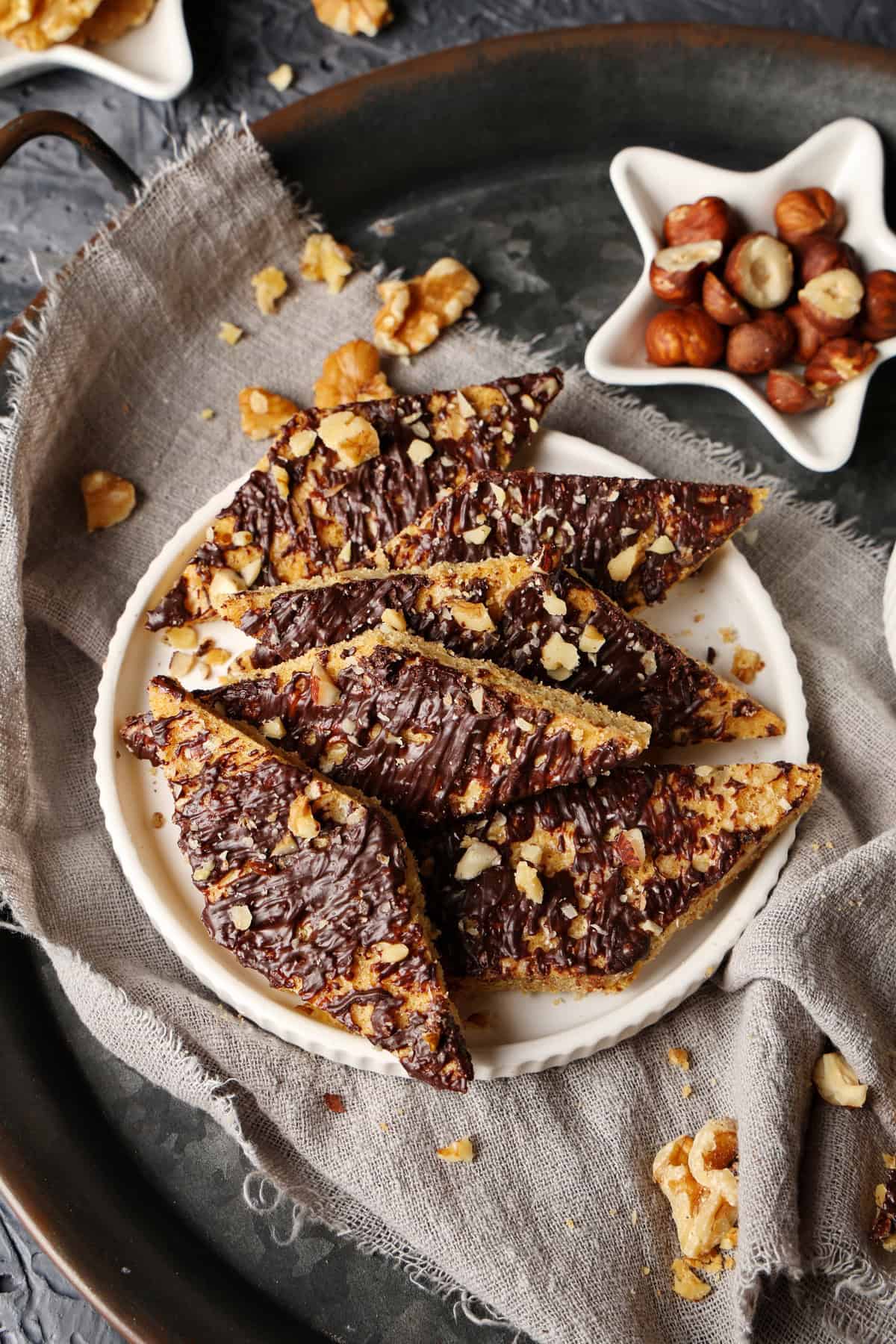 A plate of speculoos cookie bars with white chocolate. Above it, a small bowl of hazelnuts.