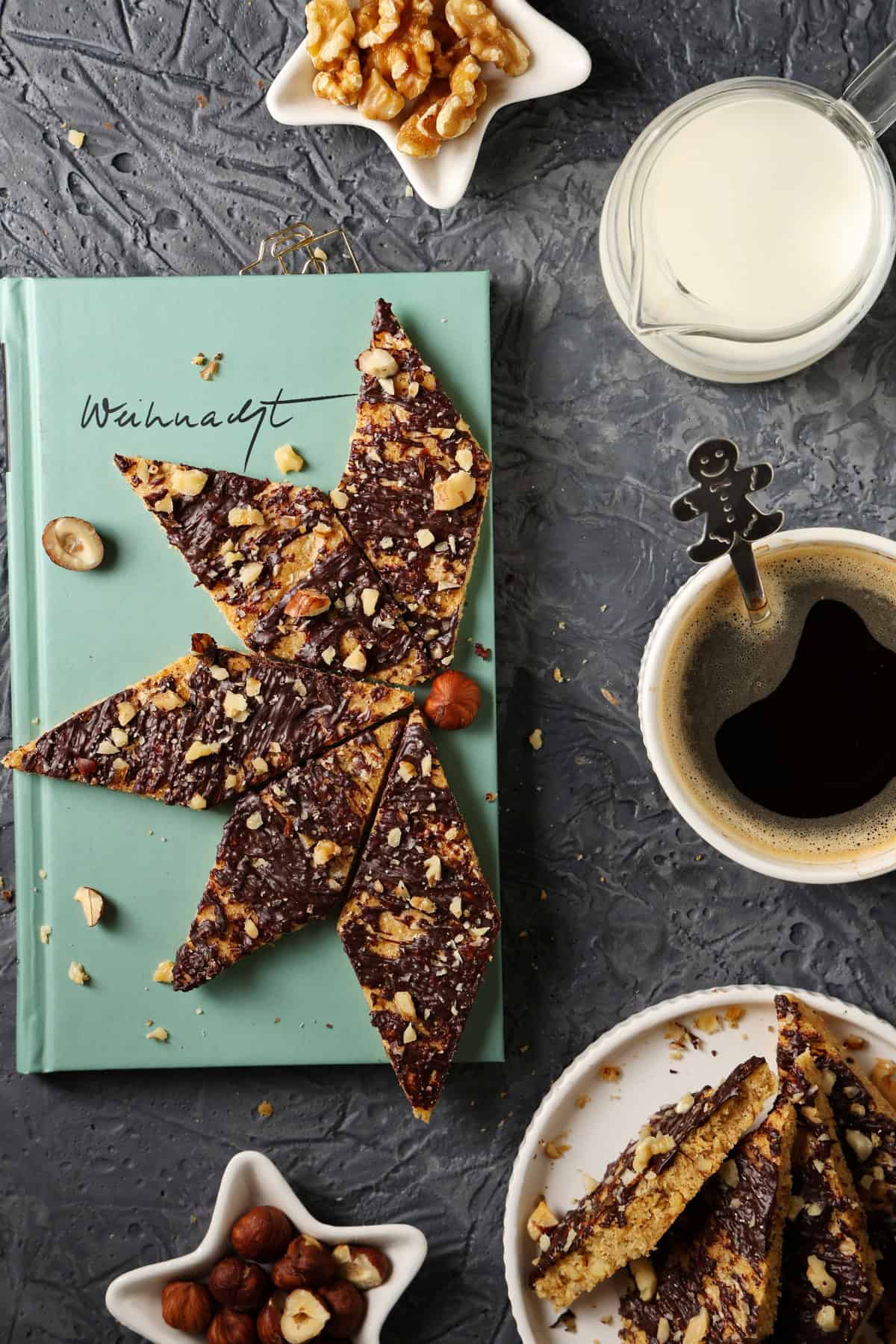 Speculoos cookie bars with white chocolate resting on a book. Next to it, a cup of coffee, milk, and nuts for decoration.