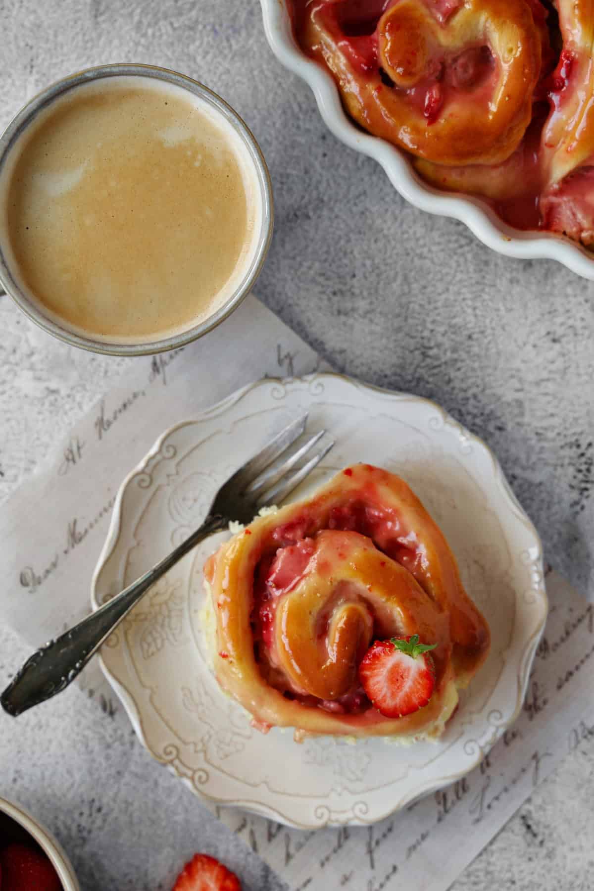 A plate with a single strawberry roll. In the background, a baking dish filled with strawberry rolls and a cup of coffee.