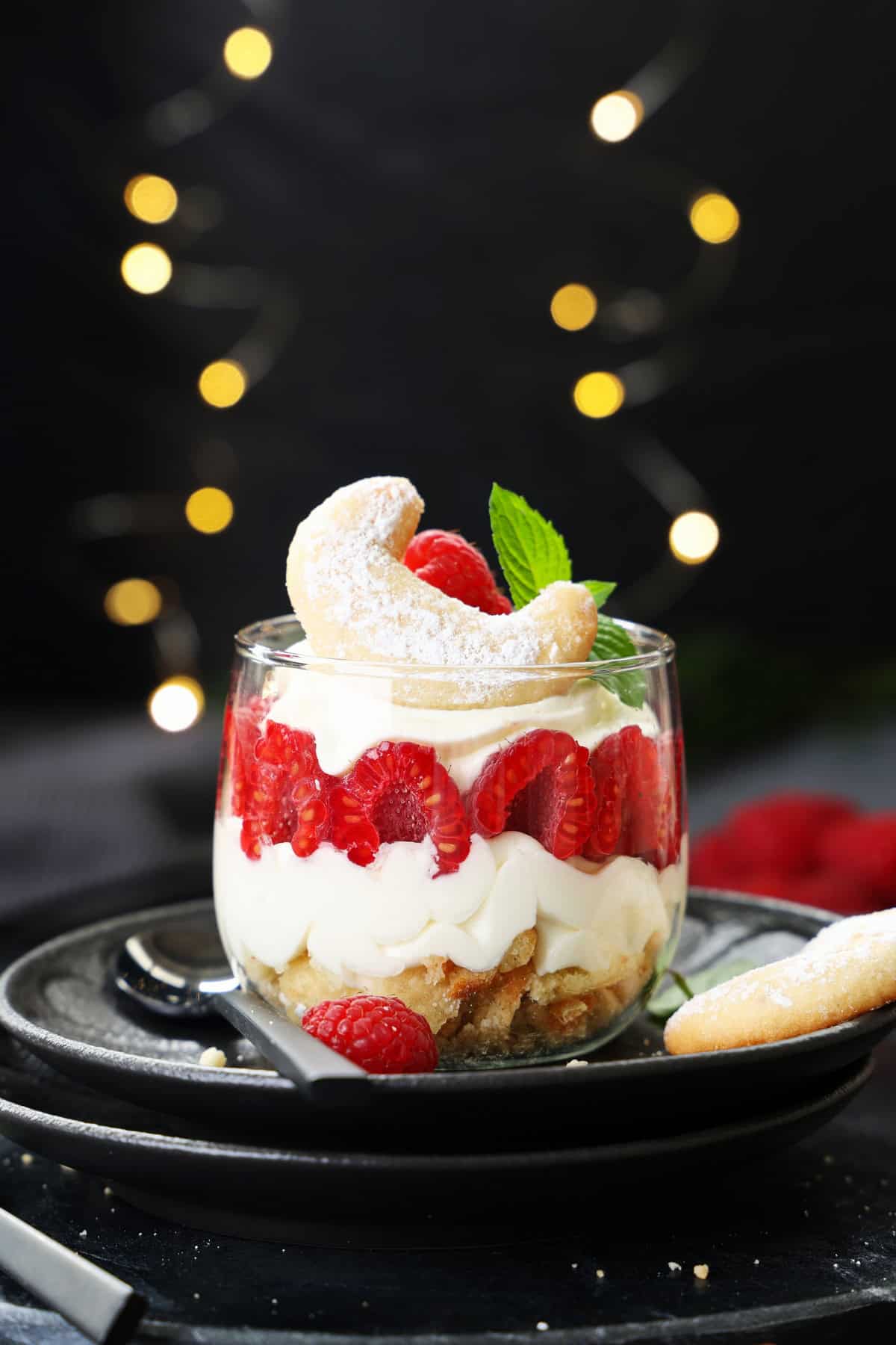 A beautifully layered vanilla crescent cookie and raspberry dessert in a glass with light cream, red berries, and a mint garnish.