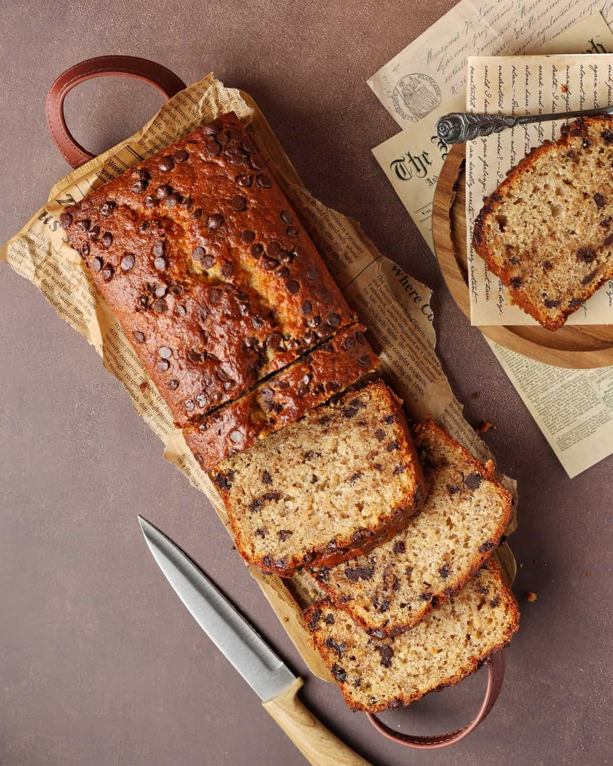 A sliced, moist banana bread with chocolate chips sits on a rustic wooden board.