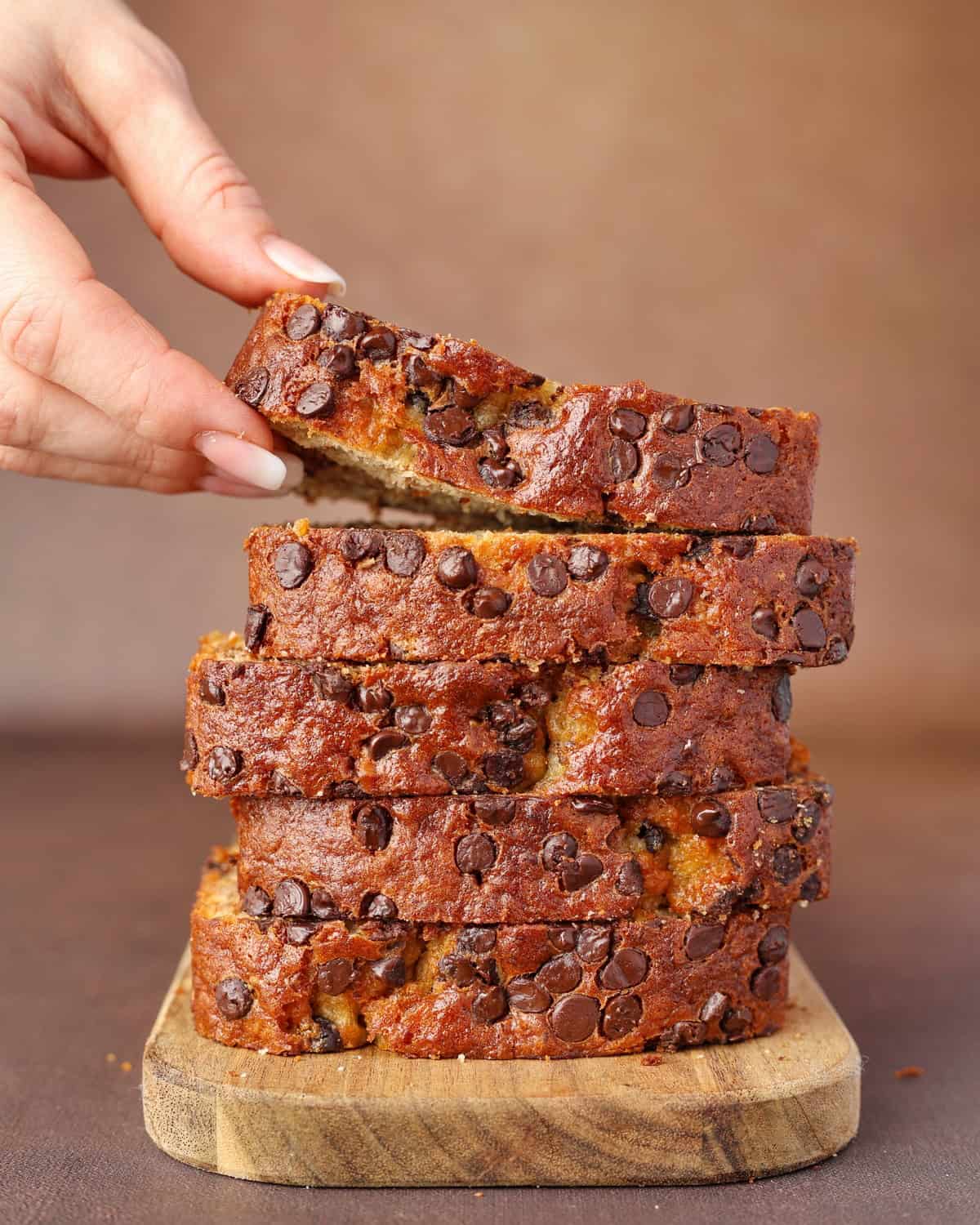 A hand places a slice of chocolate chip banana bread onto a stack of banana bread slices.