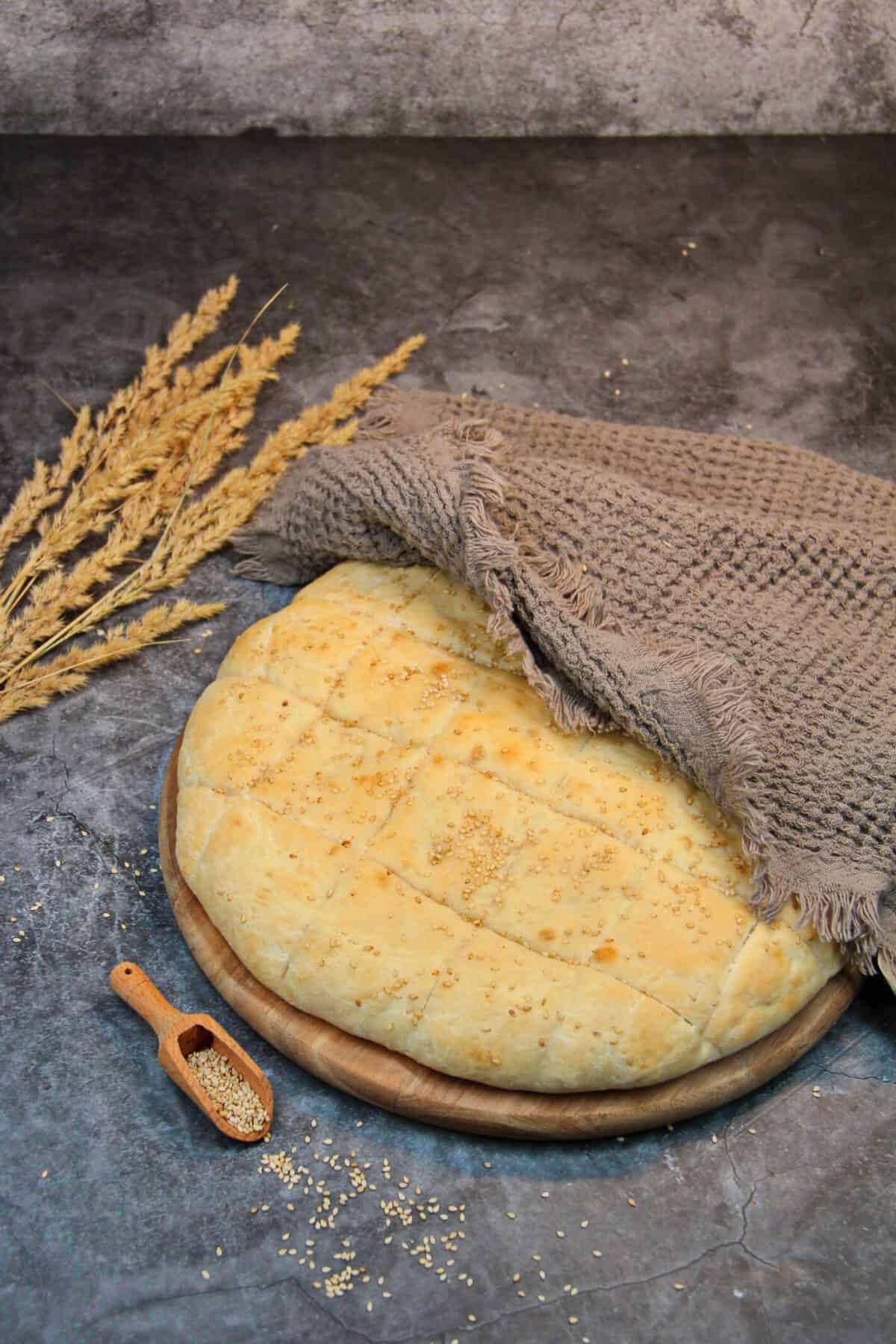 Freshly baked homemade sesame flatbread cooling under a kitchen towel on a wooden board.