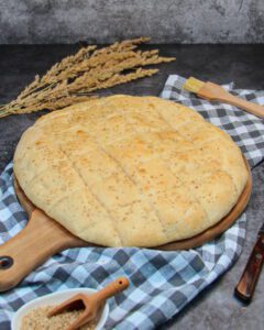 A homemade, golden-yellow flatbread with sesame seeds on a wooden board with a checkered kitchen towel.