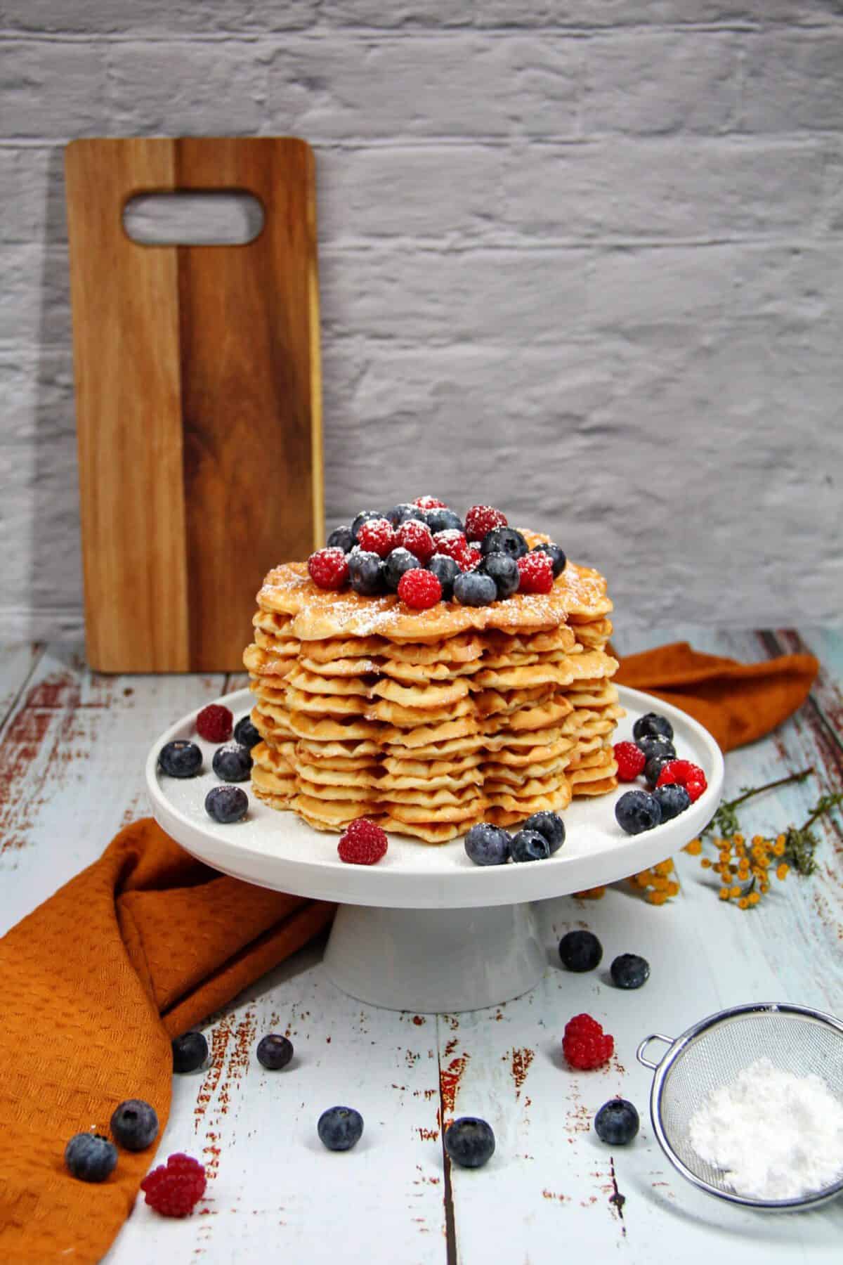 A tall stack of crispy waffles on a cake stand, topped with fresh berries and powdered sugar.