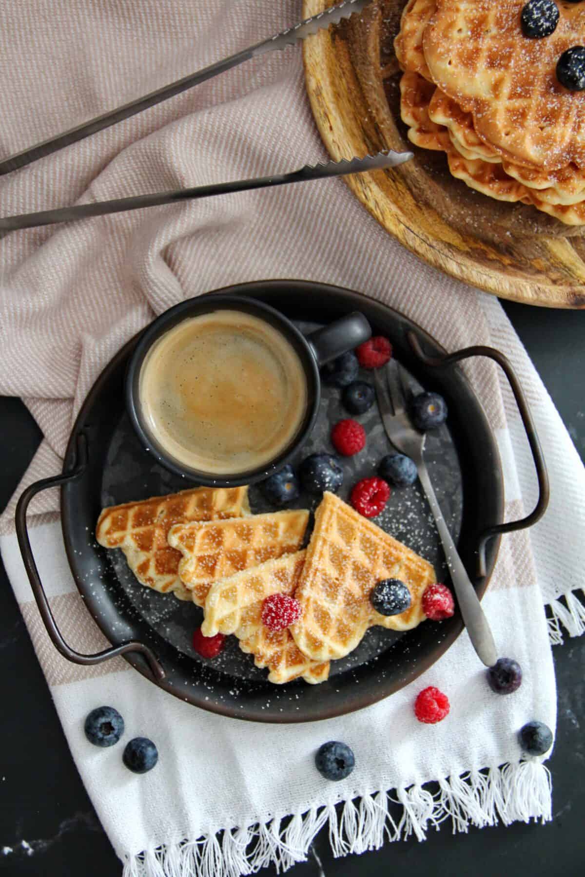 Crispy heart-shaped waffles topped with powdered sugar and fresh berries, served with coffee on a tray.