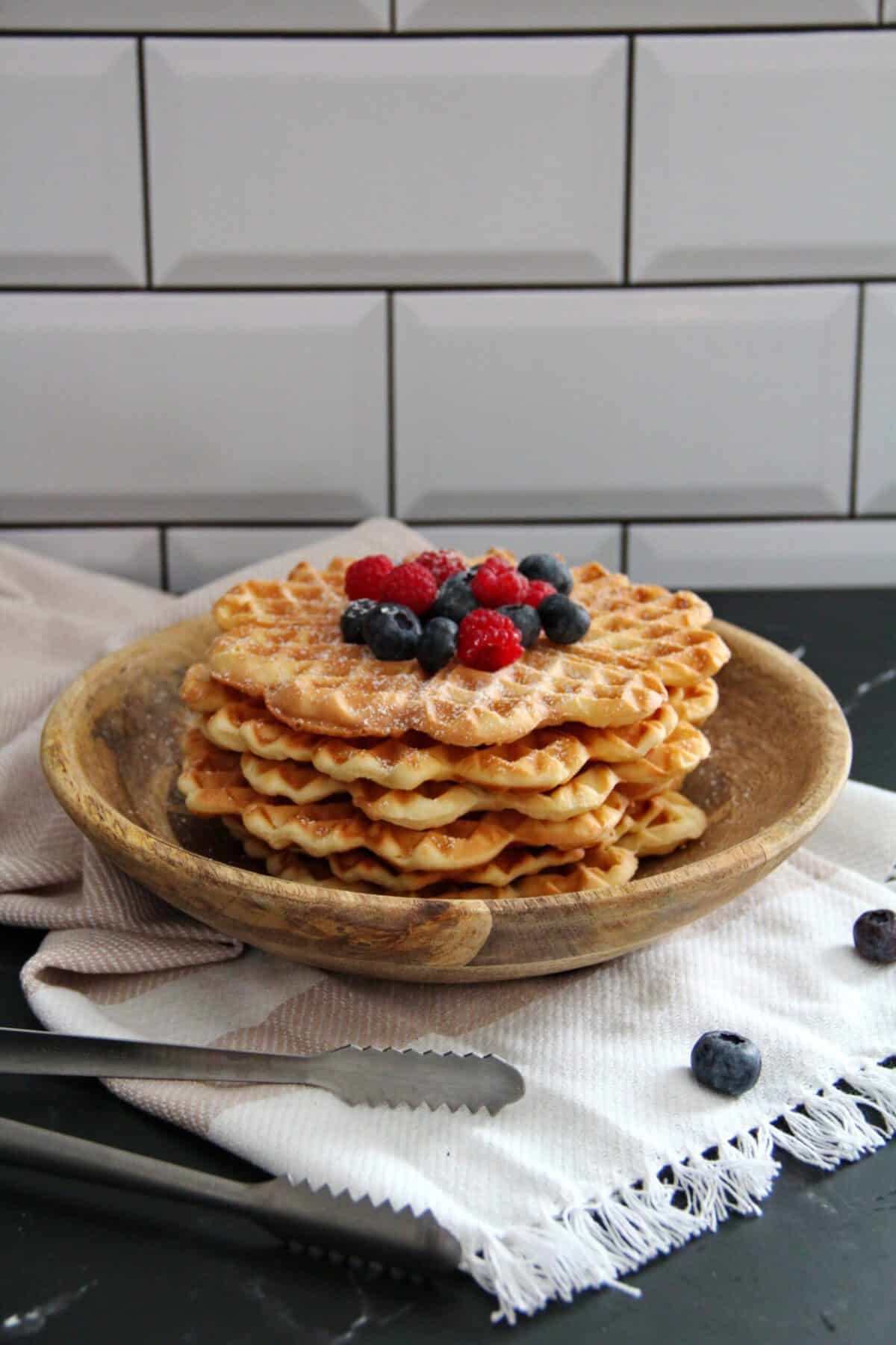 A stack of crispy waffles topped with powdered sugar, raspberries, and blueberries in a wooden bowl.