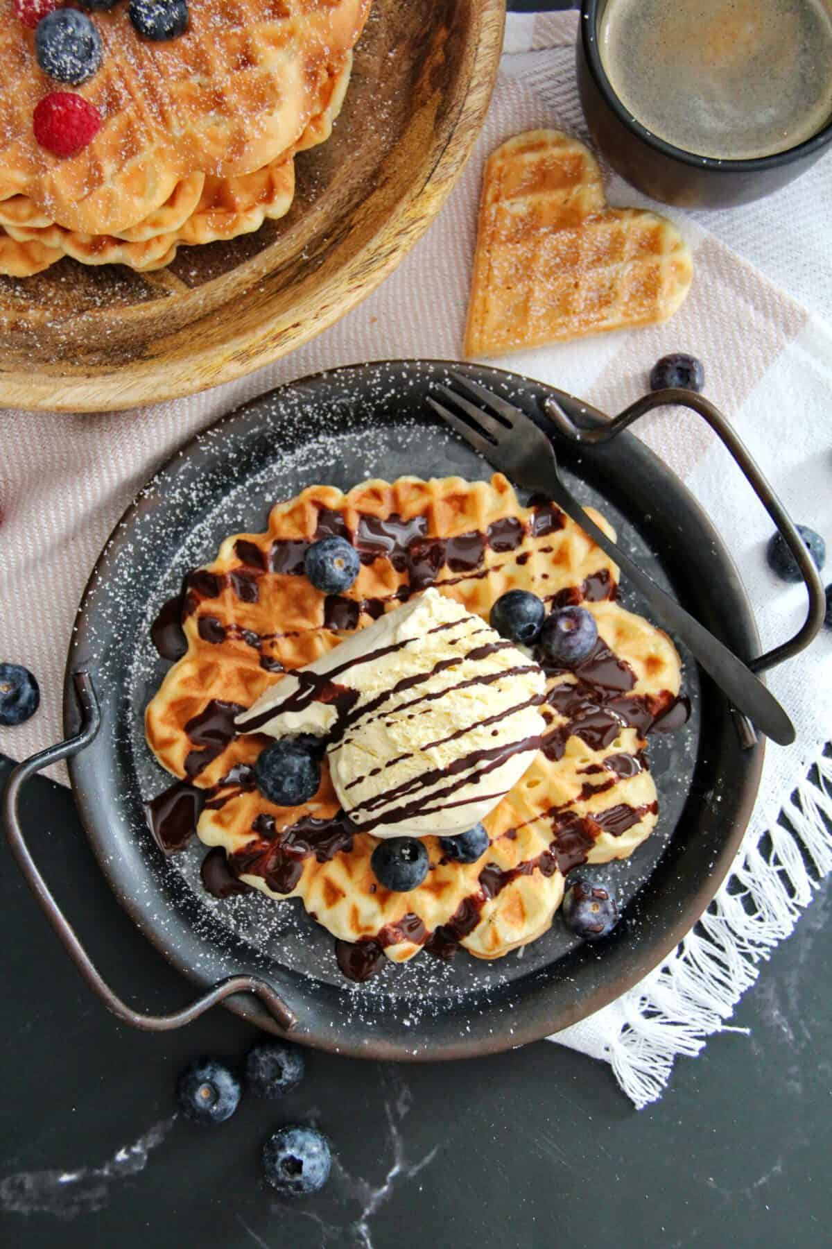 A crispy waffle served with vanilla ice cream, chocolate sauce, and fresh blueberries on a dark plate.